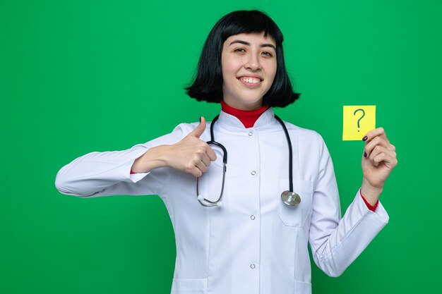 Souriante jeune jolie fille caucasienne en uniforme de médecin avec stéthoscope pouce vers le haut et tenant la note de question