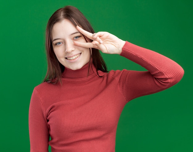 Souriante jeune jolie fille montrant le symbole v-signe près de l'œil isolé sur mur vert