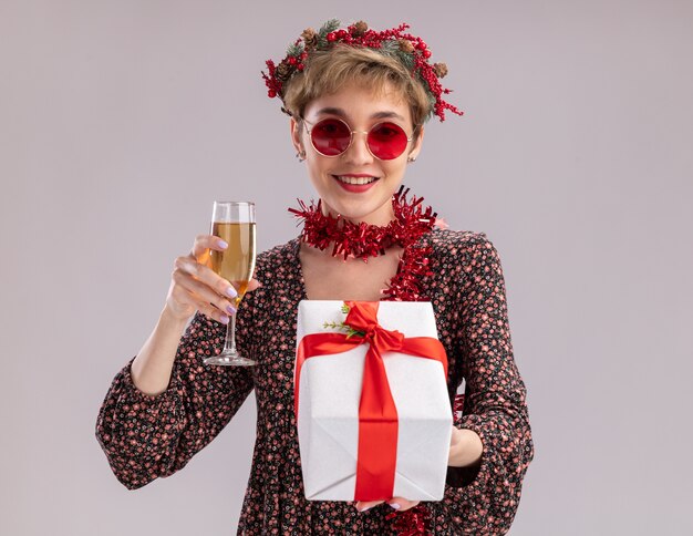 souriante jeune jolie fille portant une couronne de Noël et une guirlande de guirlandes autour du cou avec des lunettes tenant un paquet cadeau et un verre de champagne isolé sur un mur blanc avec espace de copie