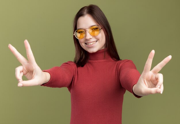 Souriante jeune jolie fille portant des lunettes de soleil faisant un signe de paix isolé sur un mur vert olive