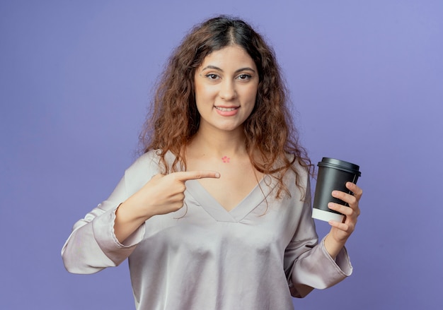 Souriante jeune jolie fille tenant et pointe une tasse de café isolé sur un mur bleu