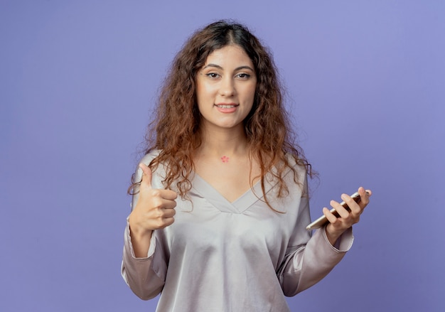 Souriante jeune jolie fille tenant le téléphone son pouce vers le haut isolé sur le mur bleu