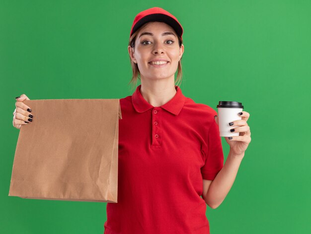 Souriante jeune jolie livreuse en uniforme détient un paquet de papier et une tasse de papier sur vert