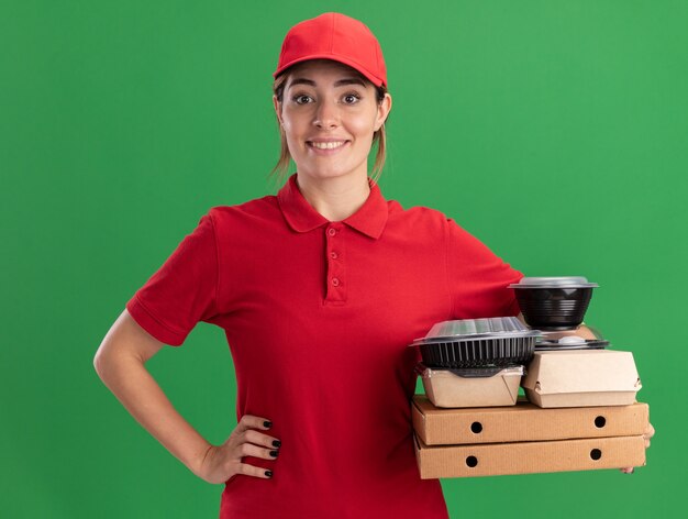 Souriante jeune jolie livreuse en uniforme met la main sur la taille et détient des emballages alimentaires en papier