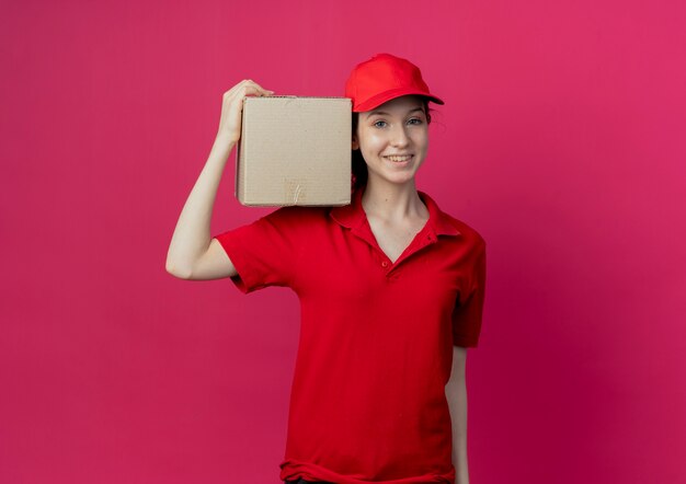 Souriante jeune jolie livreuse en uniforme rouge et cap tenant la boîte de carton sur l'épaule isolé sur fond cramoisi avec espace de copie