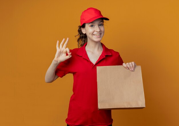 Souriante jeune jolie livreuse en uniforme rouge et capuchon montrant le paquet de papier et faisant signe ok isolé sur fond orange avec espace de copie