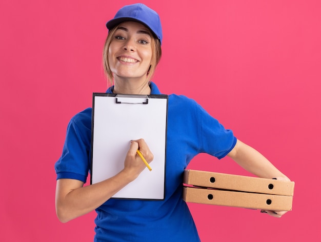 Souriante jeune jolie livreuse en uniforme tenant le presse-papiers et les boîtes de pizza sur rose