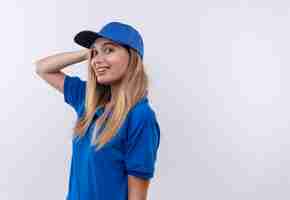 Photo gratuite souriante jeune livreuse portant l'uniforme bleu et une casquette mettant la main sur la tête isolé sur blanc