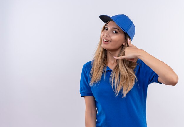 Souriante jeune livreuse portant l'uniforme bleu et une casquette montrant le geste d'appel téléphonique isolé sur un mur blanc avec espace copie