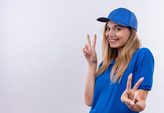 Souriante jeune livreuse portant l'uniforme bleu et une casquette montrant le geste de paix isolé sur un mur blanc avec espace de copie