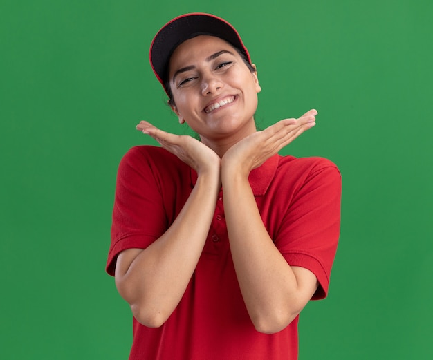 Souriante jeune livreuse portant l'uniforme et une casquette main dans la main autour du visage isolé sur un mur vert