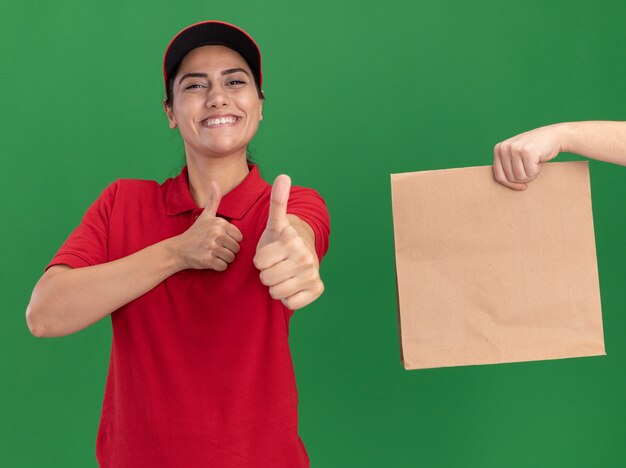 Souriante jeune livreuse portant l'uniforme et une casquette montrant le pouce vers le haut de quelqu'un qui lui donne de l'argent isolé sur un mur vert