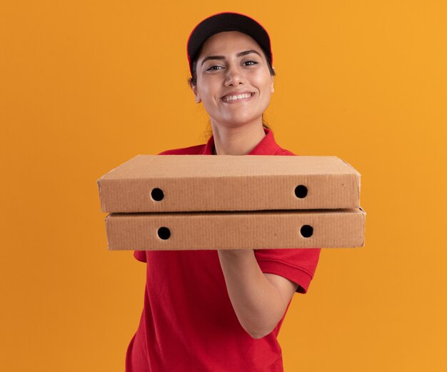 Souriante jeune livreuse portant l'uniforme et une casquette tenant des boîtes de pizza isolés sur un mur orange