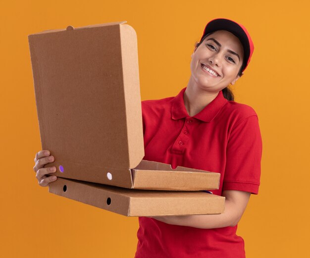 Souriante jeune livreuse portant l'uniforme et la casquette tenant et ouvrant une boîte à pizza isolée sur un mur orange