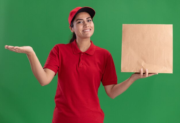 Souriante jeune livreuse portant l'uniforme et le chapeau tenant le paquet de nourriture de papier répandant la main isolée sur le mur vert