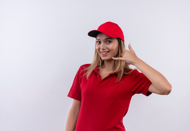 Souriante jeune livreuse portant l'uniforme rouge et une casquette montrant le geste d'appel téléphonique isolé sur blanc