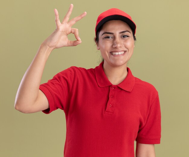 Souriante jeune livreuse en uniforme avec capuchon montrant le geste correct isolé sur mur vert olive