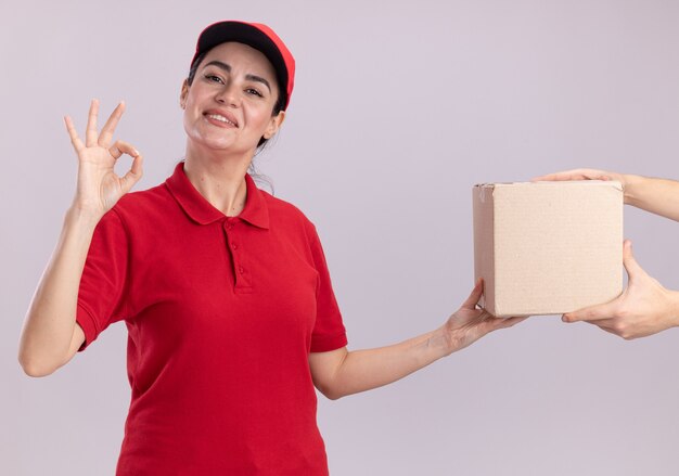 Souriante jeune livreuse en uniforme et casquette donnant une boîte à cartes au client faisant signe ok