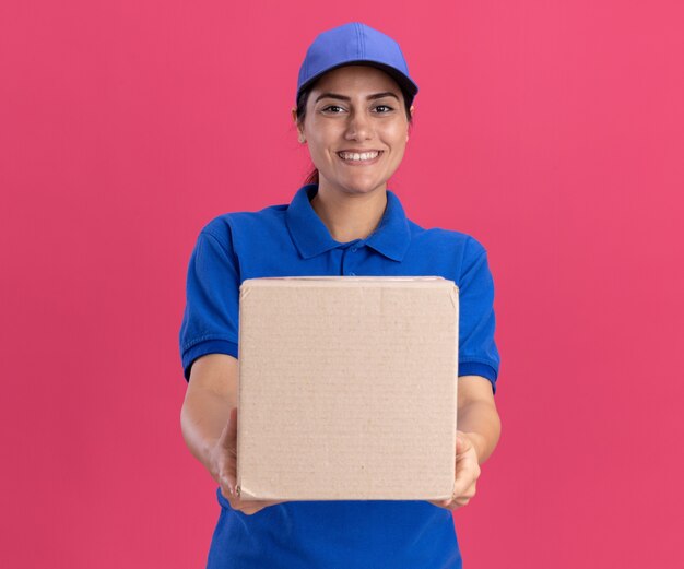 Souriante jeune livreuse en uniforme avec casquette tenant une boîte à la caméra isolée sur un mur rose