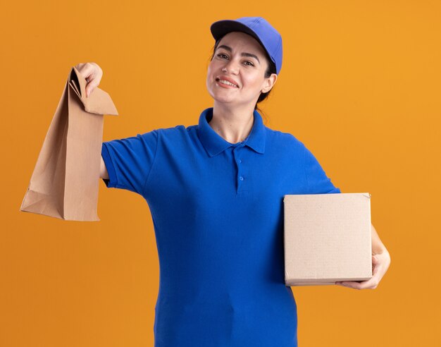 Souriante jeune livreuse en uniforme et casquette tenant une boîte à cartes et un paquet de papier isolé sur un mur orange