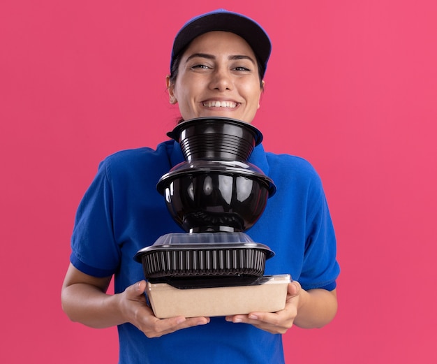 Souriante Jeune Livreuse En Uniforme Avec Casquette Tenant Des Récipients Alimentaires Isolés Sur Un Mur Rose