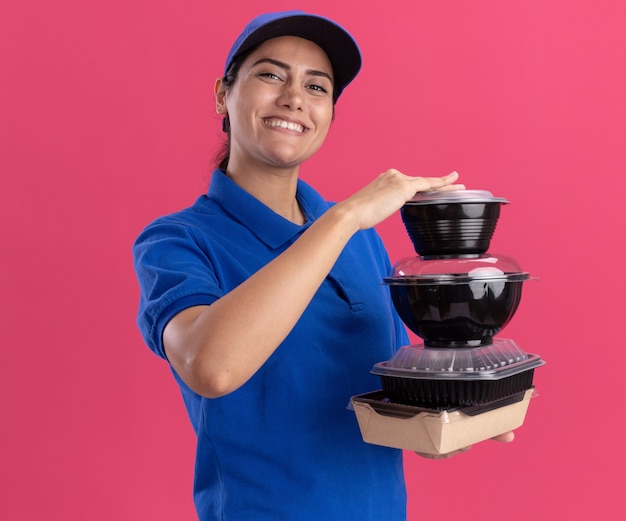 Souriante jeune livreuse en uniforme avec casquette tenant des récipients alimentaires isolés sur un mur rose