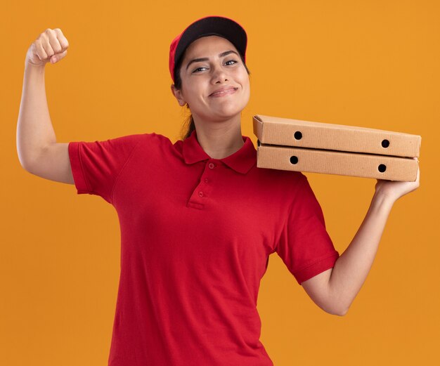 Souriante jeune livreuse en uniforme et chapeau tenant des boîtes de pizza montrant un geste fort isolé sur un mur orange