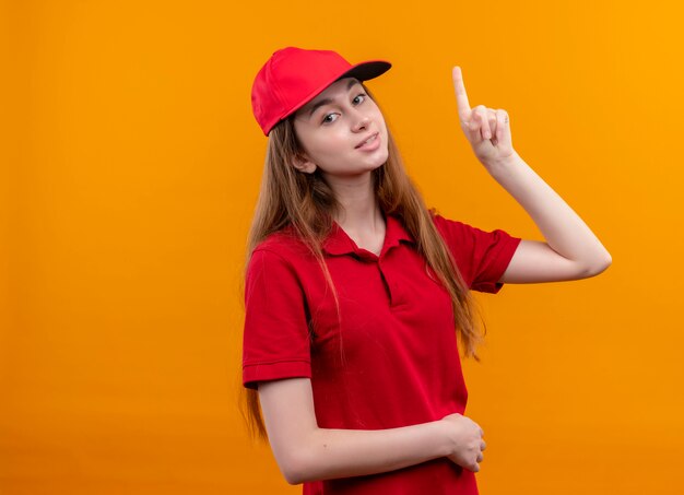 Souriante jeune livreuse en uniforme rouge pointant vers le haut et mettant la main sur le ventre sur un mur orange isolé avec espace de copie