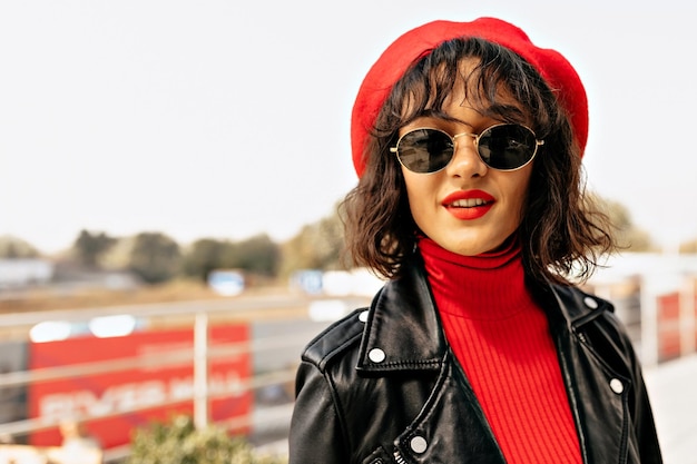 Souriante Jolie Dame Aux Lèvres Rouges Portant Une Casquette Rouge Et Une Veste Sombre Regarde La Caméra Tout En Marchant Dans La Ville Par Une Chaude Journée Ensoleillée