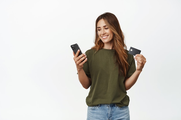 Souriante jolie femme d'âge moyen, tenant une carte de réduction de crédit, regardant l'écran du smartphone