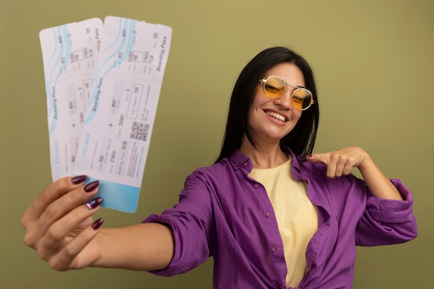 Souriante jolie femme brune à lunettes de soleil tient et regarde les billets d'avion isolés sur mur vert olive