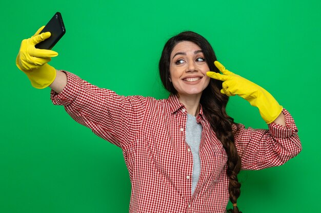 Souriante jolie femme de ménage caucasienne avec des gants en caoutchouc prenant un selfie et gesticulant le signe de la victoire