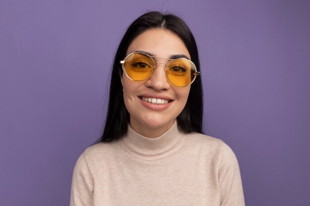 Souriante jolie fille caucasienne brune à lunettes de soleil regarde la caméra sur violet