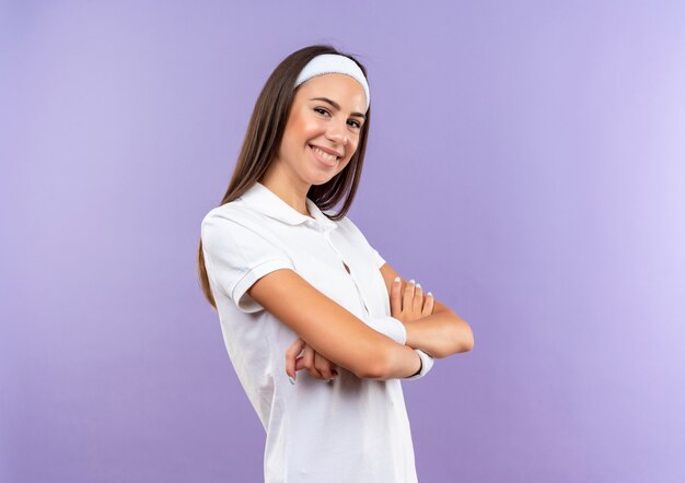 Souriante jolie fille sportive portant un bandeau et un bracelet debout avec une posture fermée isolée sur l'espace violet