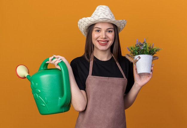 Souriante jolie jardinière caucasienne portant chapeau de jardinage tenant arrosoir et pot de fleurs isolé sur mur orange avec espace copie