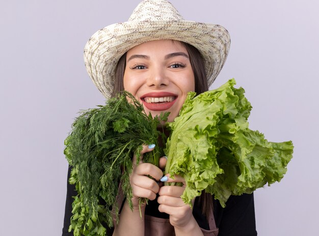 Souriante jolie jardinière caucasienne portant un chapeau de jardinage tenant un bouquet d'aneth et de salade