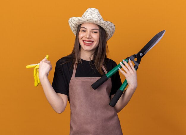 Souriante jolie jardinière caucasienne portant un chapeau de jardinage tenant des ciseaux et des gants de jardinage isolés sur un mur orange avec espace pour copie