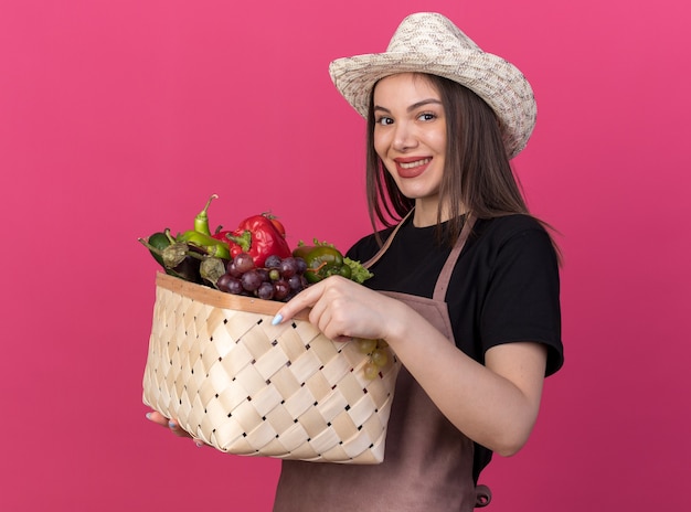 Souriante jolie jardinière caucasienne portant chapeau de jardinage tenant un panier de légumes regardant la caméra sur rose