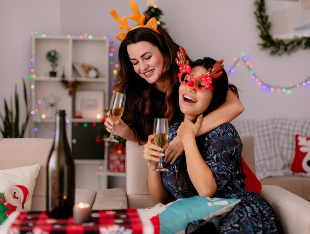 Souriante jolie jeune fille avec un bandeau de renne tient un verre de champagne et embrasse son amie avec des lunettes de renne assis sur un fauteuil et appréciant le temps de Noël à la maison
