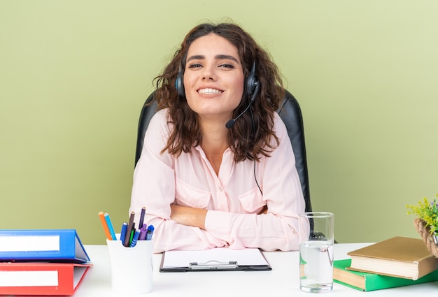 Souriante jolie opératrice de centre d'appels caucasienne sur des écouteurs assis au bureau avec des outils de bureau croisant ses bras isolés sur un mur vert