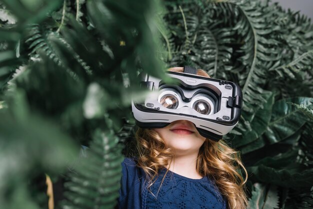 Souriante petite fille blonde utilisant un casque virtuel debout parmi la plante verte