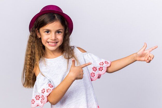 Souriante petite fille caucasienne avec chapeau de fête violet pointant sur le côté isolé sur un mur blanc avec espace copie