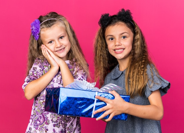 souriante petite fille caucasienne tenant une boîte-cadeau et debout avec une petite fille blonde heureuse isolée sur un mur rose avec espace de copie