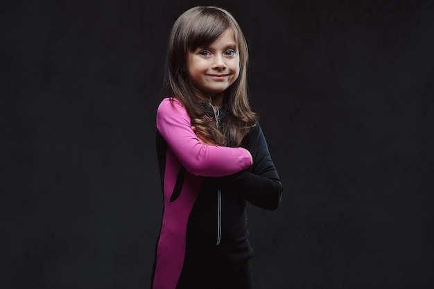 Souriante petite fille vêtue de vêtements de sport debout avec les bras croisés. Isolé sur un fond texturé sombre.