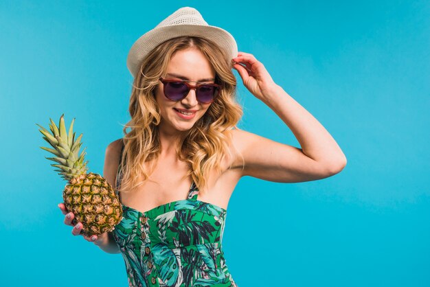 Souriante séduisante jeune femme au chapeau et des lunettes de soleil tenant l&#39;ananas