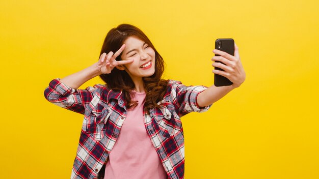 Sourire adorable femme asiatique faisant selfie photo sur smartphone avec une expression positive dans des vêtements décontractés et regardant la caméra sur le mur jaune. Heureuse adorable femme heureuse se réjouit du succès.