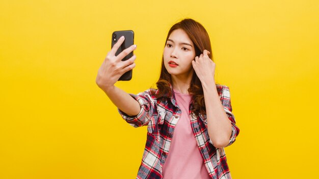 Sourire adorable femme asiatique faisant selfie photo sur smartphone avec une expression positive dans des vêtements décontractés et regardant la caméra sur le mur jaune. Heureuse adorable femme heureuse se réjouit du succès.