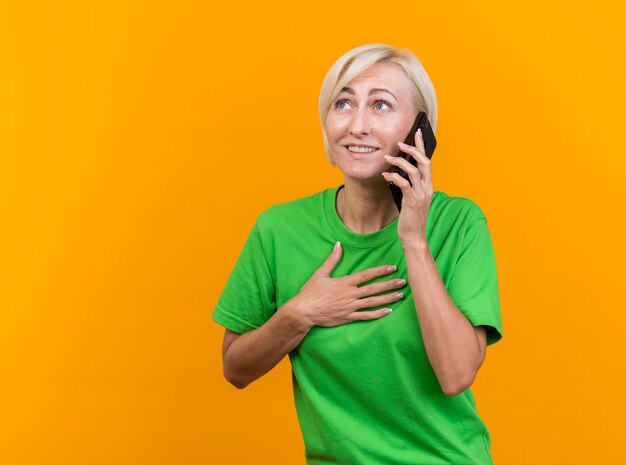 Sourire d'âge moyen blonde femme slave parlant au téléphone en gardant la main sur la poitrine à côté isolé sur mur jaune avec espace copie