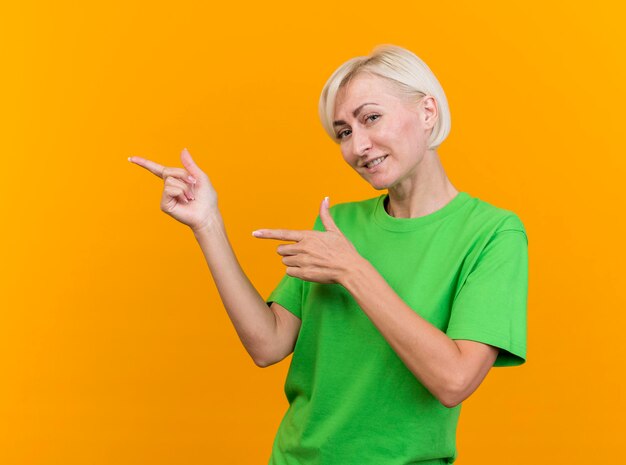 Sourire d'âge moyen blonde femme slave regardant avant pointant sur le côté isolé sur mur jaune avec espace copie