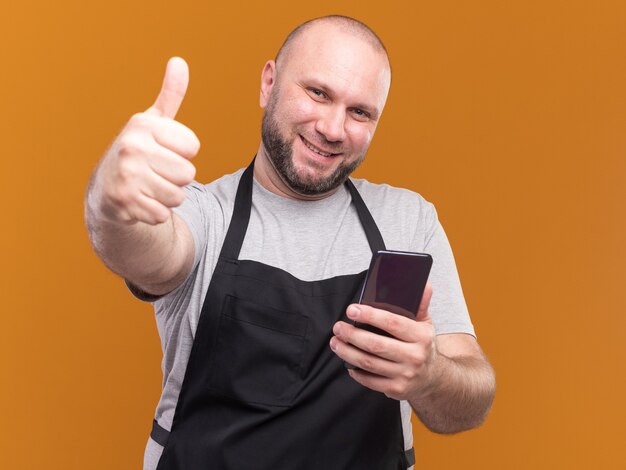 Sourire de barbier mâle d'âge moyen slave en uniforme tenant le téléphone montrant le pouce vers le haut isolé sur un mur orange